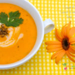 A cup of creamy carrot and lentil soup with a coriander garnish on top, with yellow backdrop and flower
