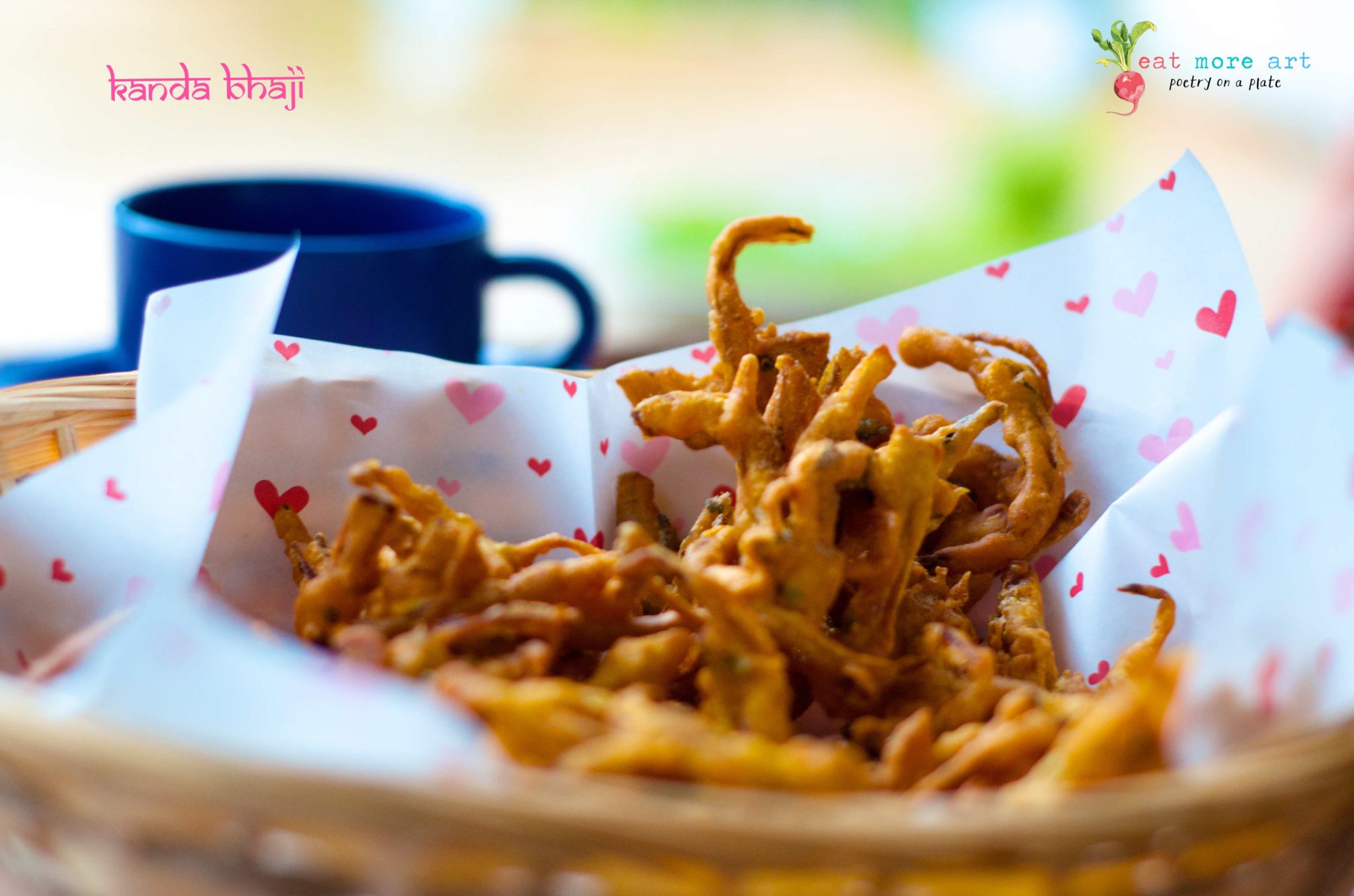 A close up of onion fritters in a basket lined with butter paper with hearts on it and a blue tea cup in background