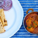 An overhead shot of Kashmir dum aloo in a copper vessel and roomali roti with onion roundels on a white plate on the side with a blue lined backdrop