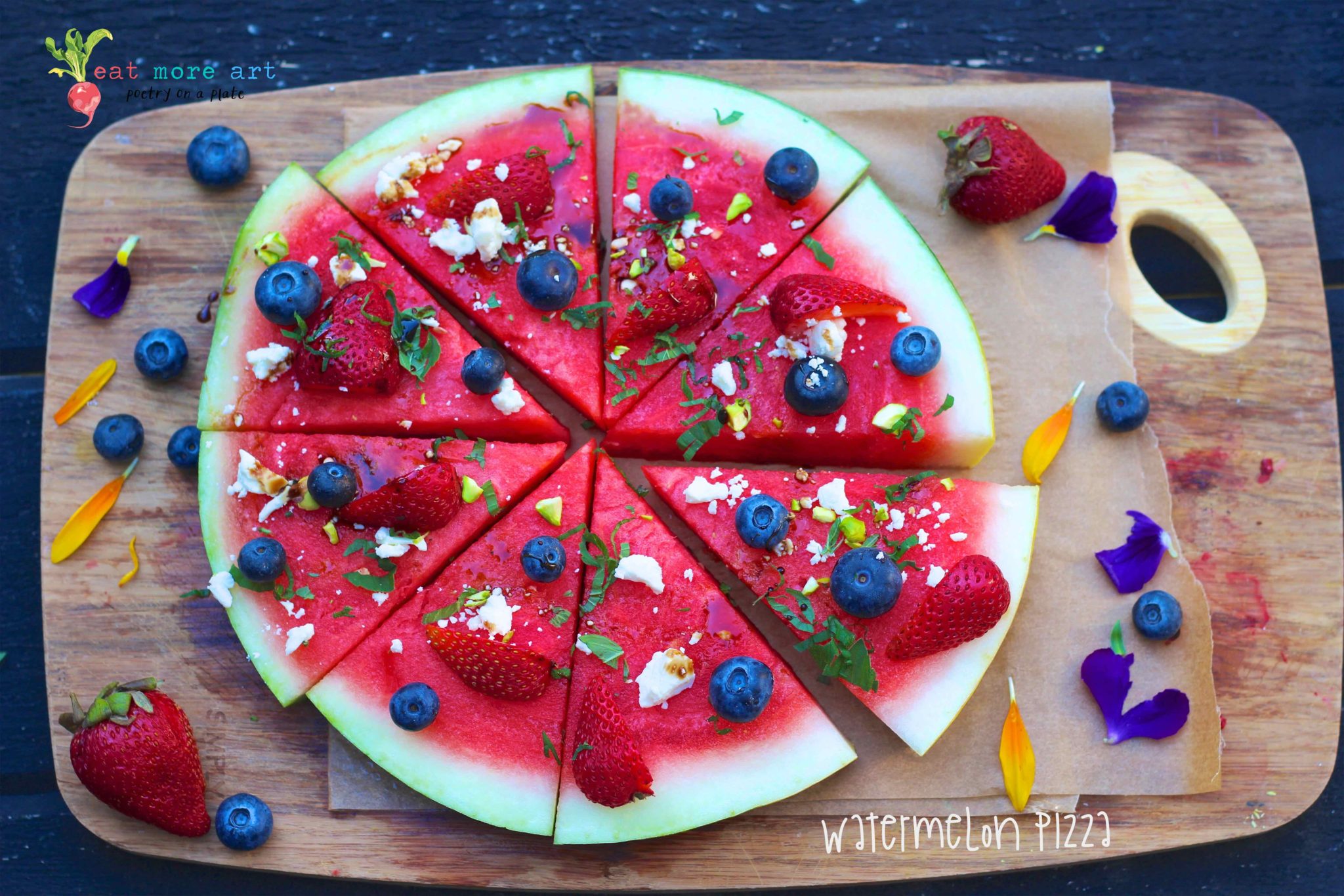 an overhead shot of Watermelon sliced like pizza topped with blueberries & strawberries feta cheese mint and balsamic vinegar on a wooden cuttingboard