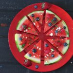 an overhead shot of Watermelon sliced like pizza topped with blueberries & strawberries feta cheese mint and balsamic vinegar on a red plate on black background