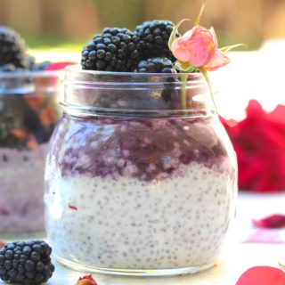 aa side shot of blackberry rose chia parfait topped with blackberries and rose petals and rose bud on a white backdrop with flower petals and blueberries scattered
