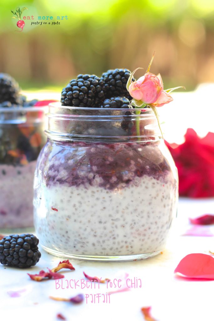 aa side shot of blackberry rose chia parfait topped with blackberries and rose petals and rose bud on a white backdrop with flower petals and blueberries scattered