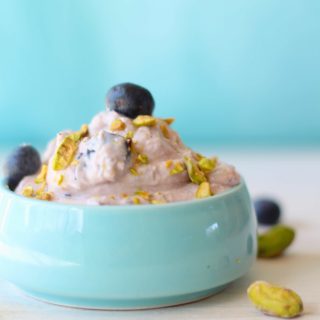 Pistachio Blueberry Shrikhand served in a baby blue bowl on a white backdrop with pistachios and blueberries scattered around