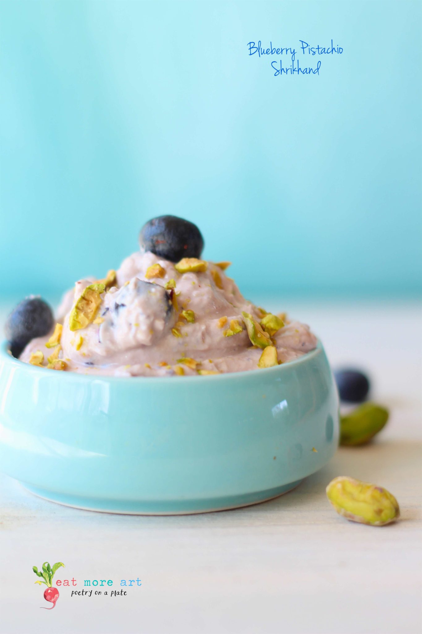 Pistachio Blueberry Shrikhand served in a baby blue bowl on a white backdrop with pistachios and blueberries scattered around